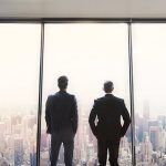 Rearview shot of two corporate businessmen looking out from a large window in their office